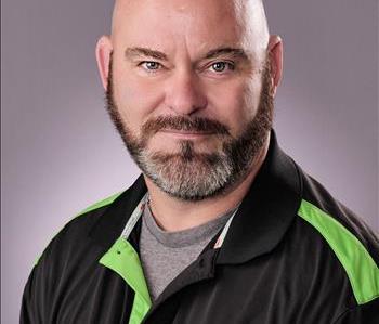 Male with buzzed dark hair and a mustache and beard. Wearing a black and green polo.